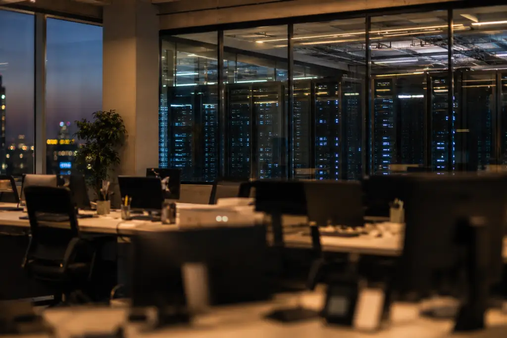 Editorial image of an office with empty desks and a glowing data center visible in the background, symbolizing tech layoffs tied to AI infrastructure investment